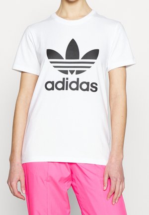 Osoba w białej koszulce Adidas z czarnym logo i jaskrawo różowych spodniach, stojąca na jednolitym tle.
