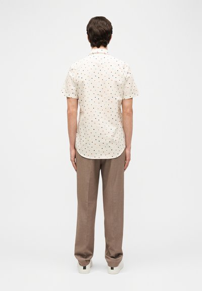 Άνδρας στέκεται γυρισμένος με την πλάτη, φορώντας λευκό κοντομάνικο πουκάμισο με μικρά πολύχρωμα τετράγωνα, καφέ παντελόνι και λευκά αθλητικά παπούτσια.