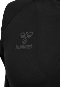 Zwarte stof met een reflecterend hummelbij-logo en tekst op de bovenrug van een kledingstuk, met zicht op textuur en stiksel-details.