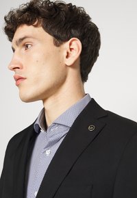 Blazer nero con tessuto testurizzato, caratterizzato da un piccolo logo metallico "MK" sulla patta sinistra. Sotto c'è una camicia con bottoni a micro-pattere blu.