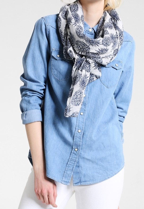 Personne portant une chemise en denim bleu clair avec les manches retroussées et une écharpe blanche imprimée d’ananas et de feuilles.