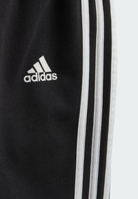 Czarne spodnie sportowe z białymi paskami po bokach, z małym białym logo adidas na udzie. Wykonane z miękkiego, teksturowanego materiału.