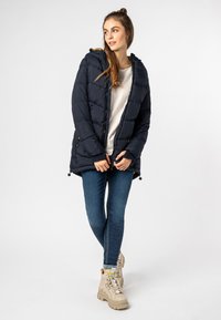 Vrouw in een navy pufferjack, witte shirt, skinny jeans en beige chunky laarzen, staand met gekruiste benen en kijkend naar rechts.