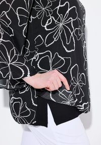 Blusa nera con motivo floreale bianco, materiale trasparente, vestibilità ampia, spacchi laterali, indossata sopra una parte inferiore nera, che mette in mostra dettagli di design delicati.