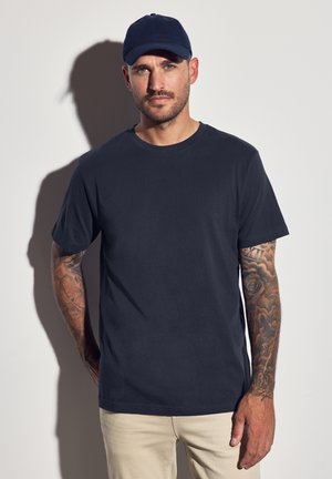 Camiseta de algodón de cuello redondo en color marino, de manga corta, con un corte relajado. Tatuajes visibles en el brazo del modelo. El fondo claro realza el contraste.