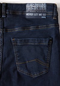 Donkerblauwe denim jeans met oranje stiksel, een leren label met de tekst "DENIM NEVER LET ME GO CECIL" en een achterzak met een klassieke snit.