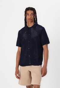 Camisa de punto de manga corta de color azul marino con un diseño de tejido abierto, frente con botones y cuello acanalado, combinada con pantalones cortos de color beige claro.