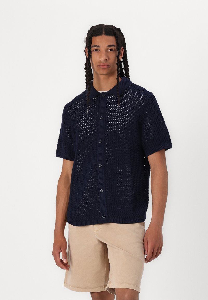 Camisa de punto de manga corta de color azul marino con un diseño de tejido abierto, frente con botones y cuello acanalado, combinada con pantalones cortos de color beige claro.
