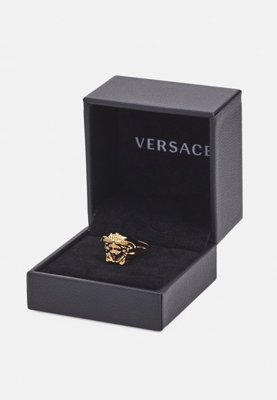 Zlatý prsteň s dizajnom leva, vystavený v čiernej koženkovej krabičke s plyšovým interiérom. Viditeľné meno značky "Versace".