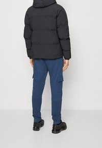 Person klädd i svart puffjacka, blå cargobyxor med sidofickor och svarta sneakers, stående mot en enfärgad vit bakgrund.