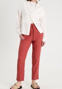 Femme portant un pantalon à cordon de serrage couleur rouille, une chemise blanche par-dessus un haut blanc, et des sandales beiges cloutées, debout devant un fond uni.
