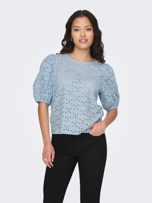 Femme portant un chemisier bleu clair à manches bouffantes en broderie anglaise et un pantalon noir, debout avec une main dans la poche sur un fond uni.