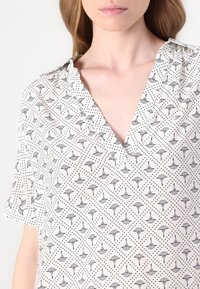 Blouse légère à manches courtes en crème avec un décolleté en V. Présente un motif de éventail noir et un motif à pois sur l'ensemble. Texture de tissu douce.
