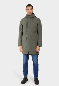 Vodoodporna parka olivno zelene barve s kapuco, sprednjim zadrgo in žepki na zaskočke, nošena preko modrih kavbojk in črnih škornjev, z ravnim krojem.