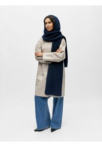 Manteau beige mi-long avec manches longues, associé à un jean large bleu et une écharpe en tricot bleu foncé. Des chaussures noires à bout pointu complètent la tenue.