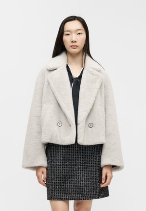 Woman wearing a cropped, light gray faux fur coat over a black blouse and a black and white patterned skirt, standing against a plain background.