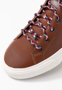 Brązowe skórzane trampki z kontrastującą białą gumową podeszwą, z kolorowymi wzorzystymi sznurówkami i czarnymi oczkami dla pewnego zapięcia.