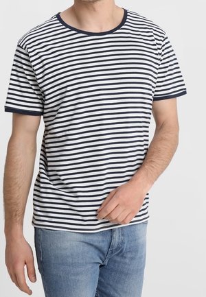 Uomo che indossa una t-shirt bianca a maniche corte con strisce orizzontali nere e jeans blu su uno sfondo grigio chiaro uniforme.