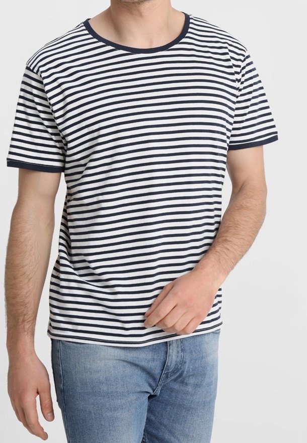 Homme portant un t-shirt blanc à manches courtes avec des rayures horizontales noires et un jean bleu sur un fond gris clair uni.
