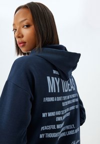 Sudadera azul marino hecha de tejido suave, con un gran estampado gráfico en texto gris claro en la espalda, y capucha con cordones.