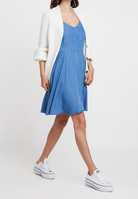 Jeansklänning i ljusblått med A-linjeform, med V-ringning och nederkant med fransar, i kombination med en vit cardigan och vita sneakers.