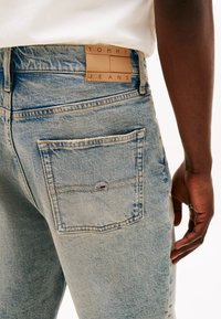 Light blue denim jeans feature a back pocket with a small, branded logo. Distressed texture and straight cut design are visible.