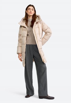 Beige pufferjack met een capuchon, ritssluiting en zijzakken. G gedragen over een lichtgrijze trui en losse donkere grijze broek.