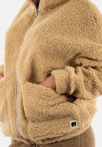 Giacca in pile beige con zip frontale, texture morbida, tasche laterali e un'etichetta con logo in basso a sinistra. Maniche lunghe e collo alto.