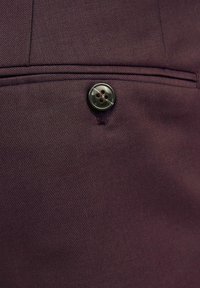 Tissu bordeaux présentant un tissage texturé, avec un bouton noir cousu au bord d'une poche, mettant en valeur une finition soignée.