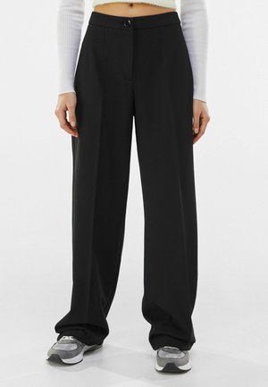 Schwarze, weit geschnittene Hose mit vorderem Knopf und Reißverschluss, kombiniert mit grauen Sneakern und einem weißen, langärmeligen, kurzen Oberteil.