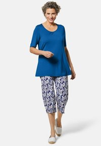 Blaues kurzärmliges Oberteil kombiniert mit gemusterten, knöchellangen Hosen, die weiße Blumenmuster auf einem dunkelblauen Hintergrund zeigen. Helle Schuhe runden das Outfit ab.