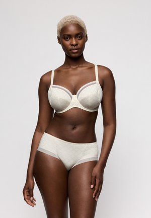Vrouw met kort blond haar die een bijpassende witte beha en slipje met subtiel patroon draagt, poserend tegen een effen lichte achtergrond.