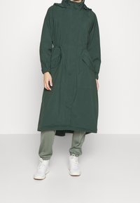 Dlouhý, tmavozelený lehký nepromokavý plášť s vysokým límcem, elastickými manžetami a dvěma předními kapsami. Nosit přes světle zelené kalhoty a bílé tenisky.