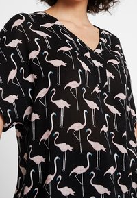 Schwarzes Shirt mit V-Ausschnitt, verziert mit einem Muster von rosa Flamingos mit blauen Akzenten. Leichter Stoff mit lockerer, lässiger Passform.