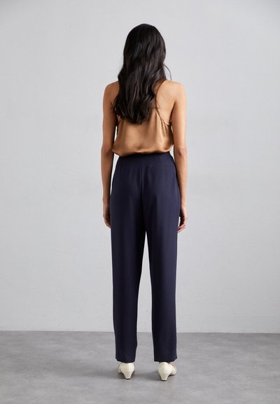 Satin brown sleeveless top paired with navy trousers. Trousers have a straight cut and a back pocket, complemented by white heeled shoes.