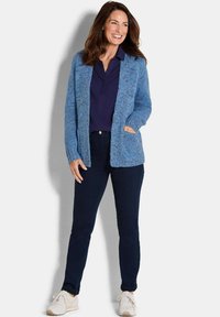 Frau lächelt, trägt eine blaue Strickjacke über einem dunkelblauen Shirt, dunkle Jeans und weiße Turnschuhe und steht vor einem schlichten hellen Hintergrund.