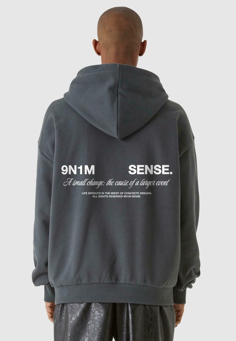 Personne portant un sweat à capuche gris foncé avec un texte blanc au dos indiquant « 9N1M SENSE. Un petit changement : la cause d’un événement plus important ».