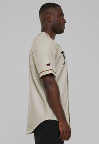 Maglietta da baseball beige con texture perforata, caratterizzata da scritte nere e dettagli marroni sul colletto e sulle maniche. Vista laterale mostrata.