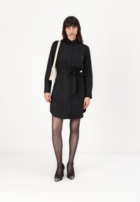 Svart skjortklänning med krage, knappframsida och tygband i midjan. Styllt med genomskinliga strumpbyxor och svarta högklackade skor.