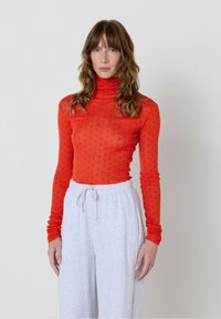 Sieviete ar viļņainiem matiem valkā pieguļošu oranžu turtleneckreklu ar zilām punktītēm un brīvus gaiši pelēkus sporta bikses uz vienkārša fona.