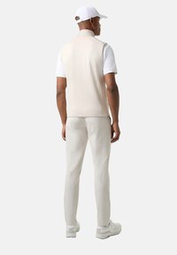 Hellbeige ärmellose Weste über einem weißen kurzärmeligen Hemd, kombiniert mit hellbeigen Hosen und weißen Sneakers, von hinten betrachtet.