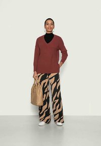 Sweater bordeaux com decote em V, camisa preta de gola alta por baixo, combinado com calças largas em padrão bege e preto. Segura uma bolsa bege; sapatos brancos.