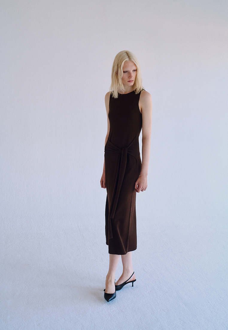 Femme aux cheveux blonds portant une robe midi noire sans manches avec un détail noué à l'avant et des escarpins slingback noirs à bout pointu, debout sur un sol clair.