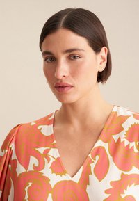Blusa con motivo floreale caratterizzata da una scollatura a V, maniche corte e disegni ondulati rosa e giallo su uno sfondo bianco.