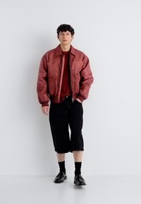 Νεαρό άτομο που φοράει κόκκινο μπουφάν τύπου bomber, κόκκινο πουκάμισο, μαύρα σορτς μέχρι το γόνατο, μαύρες κάλτσες και μαύρα παπούτσια, στέκεται μπροστά σε λευκό φόντο.