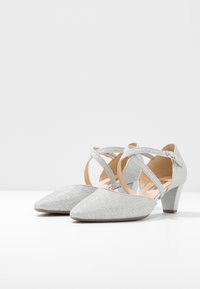 Scarpe a basso tacco in glitter argento con punta appuntita. Presentano un cinturino alla caviglia incrociato e una tomaia testurizzata. Interno liscio.