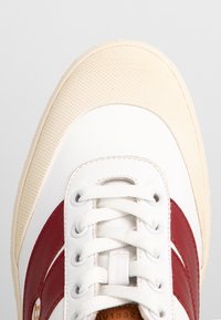 Baskets blanches avec bande latérale rouge, lacets blancs et bout beige texturé, vues du dessus sur un fond gris clair.