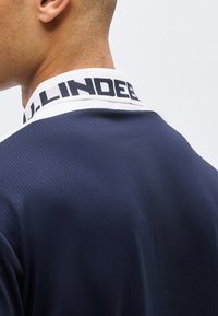 Άνδρας που φοράει μπλε σκούρα μπλούζα με λευκό γιακά και τολμηρό μαύρο κείμενο κατά μήκος της άκρης, απεικονιζόμενος από πίσω.