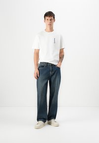 Camiseta blanca de algodón con un pequeño logo azul, combinada con unos vaqueros oscuros de corte holgado y zapatillas color crema, de pie contra un fondo liso.