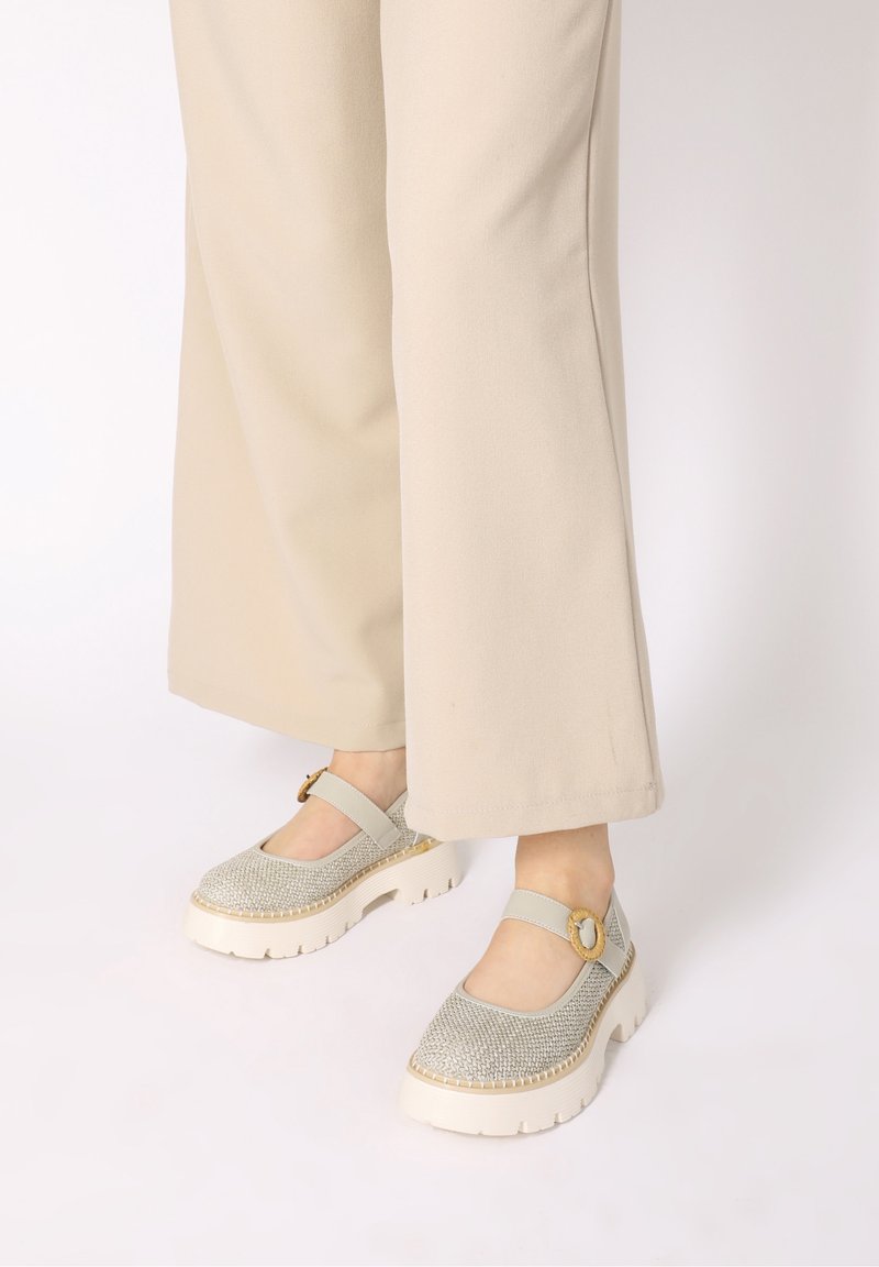 Pieds chaussés de chaussures Mary Jane tissées beige clair avec semelles blanches épaisses et brides à boucle, associées à un pantalon beige à larges jambes.